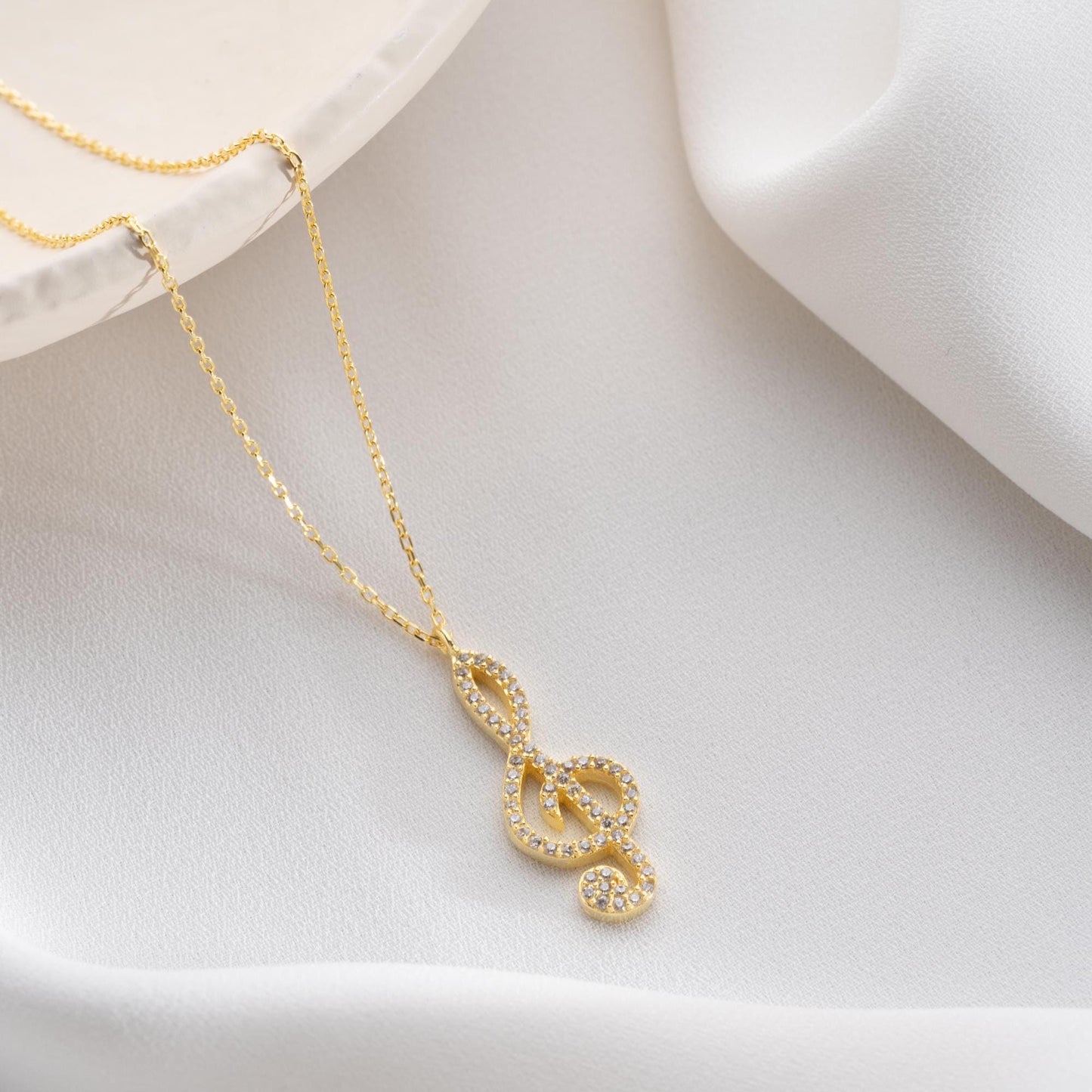 Music note necklace with cubic zirconia stones, available in 925 sterling silver and 14K gold plated options, displayed on a white satin background.
