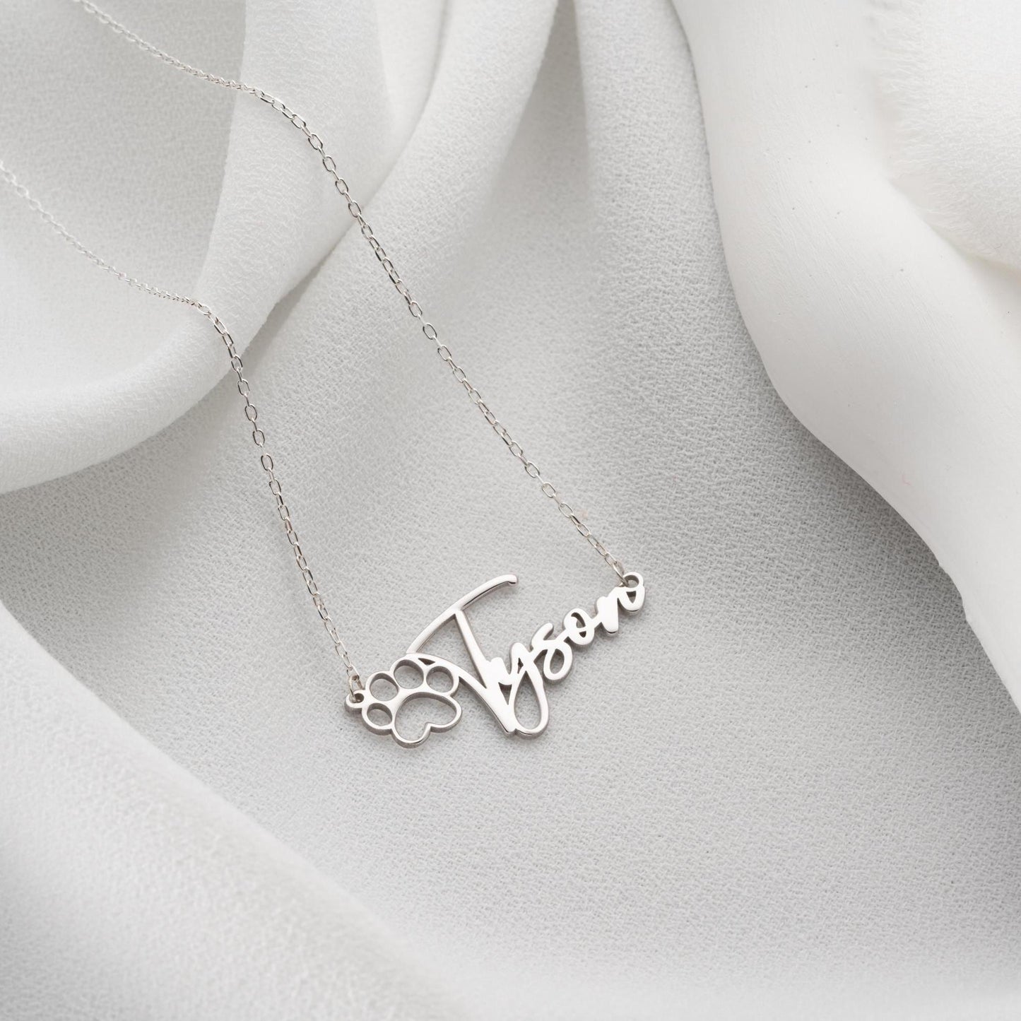 Custom pet name necklace with a paw print design in 925 sterling silver and 14k gold plated options, perfect for pet lovers. A personalized, handmade gift displayed on a soft fabric background.