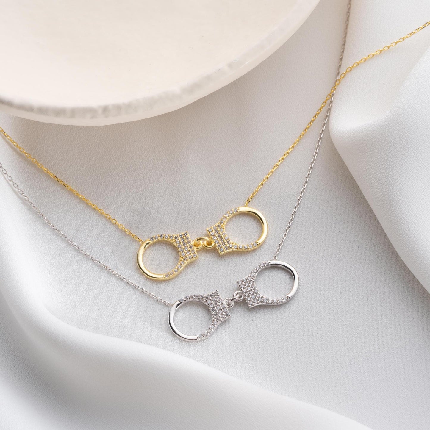 Handcuff necklace with cubic zirconia stones, available in 925 sterling silver and 14K gold plated options, displayed on a white satin background.