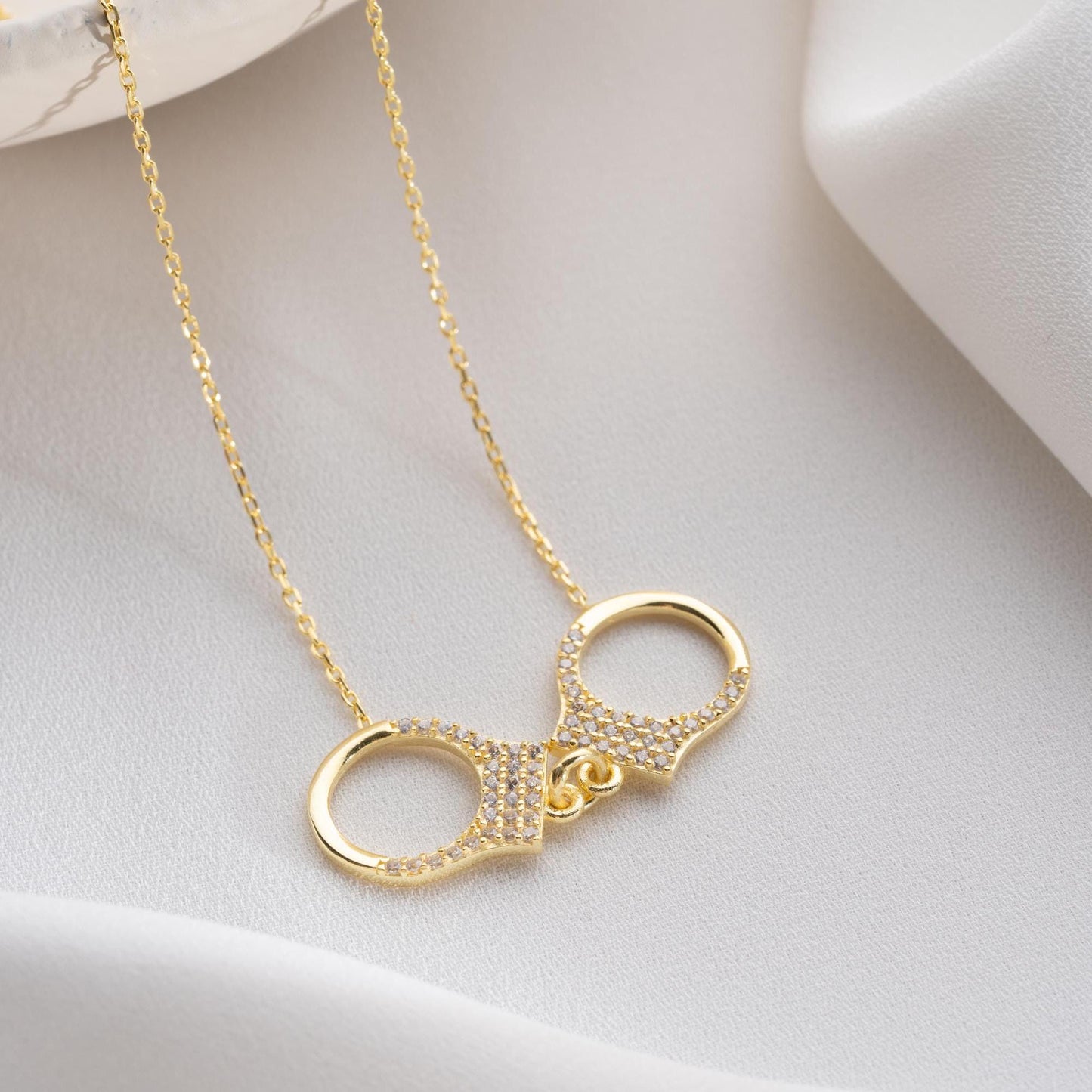 Handcuff necklace with cubic zirconia stones, available in 925 sterling silver and 14K gold plated options, displayed on a white satin background.
