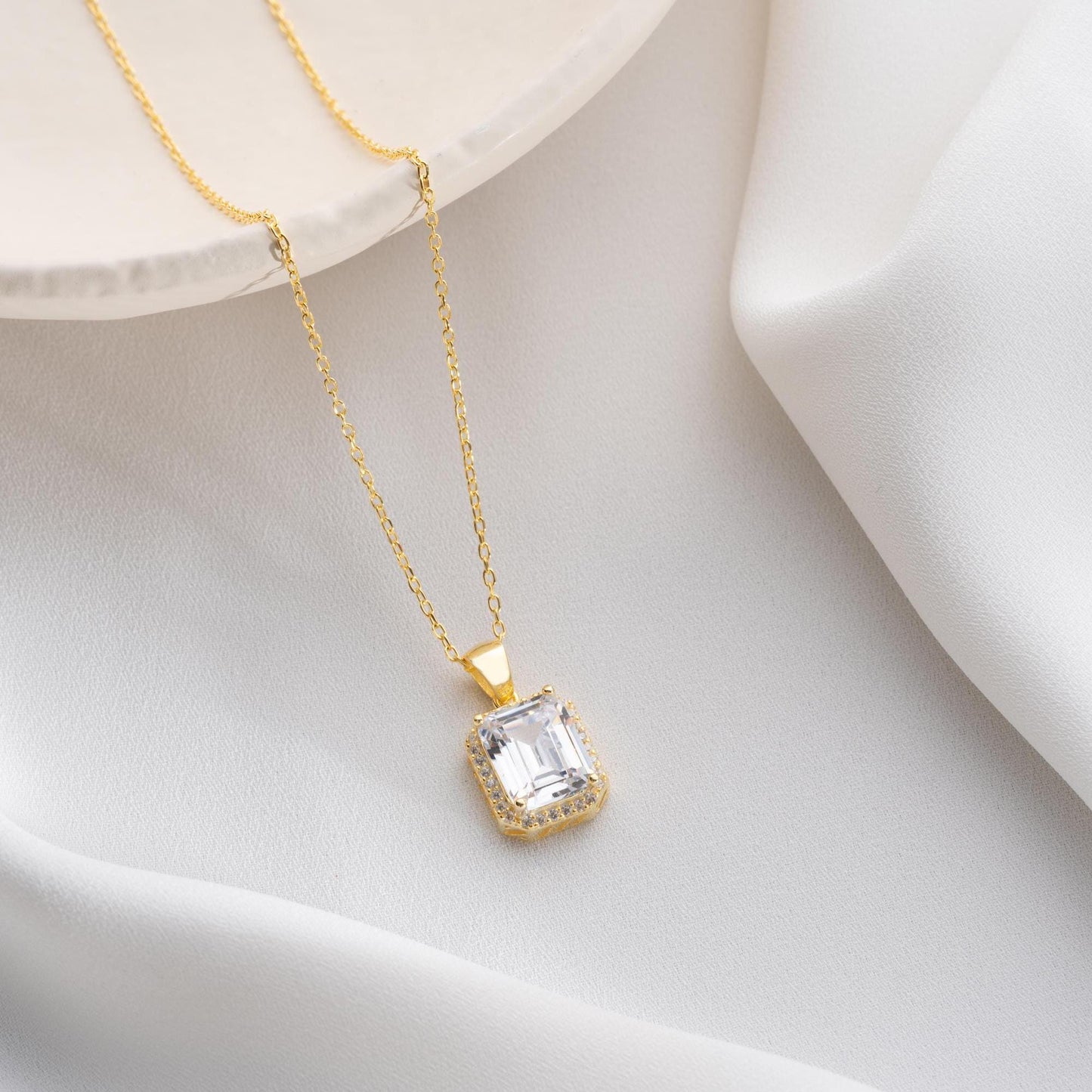Emerald-cut pendant necklace with cubic zirconia stones, available in 925 sterling silver and 14K gold plated options, displayed on a white satin background.