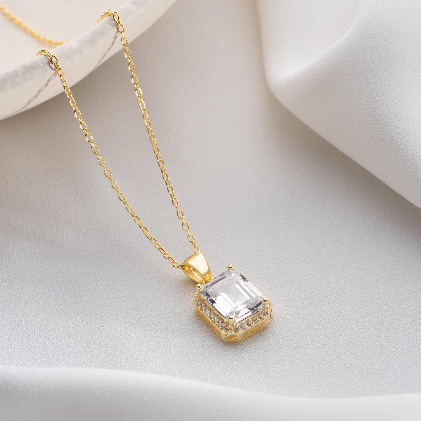 Emerald-cut pendant necklace with cubic zirconia stones, available in 925 sterling silver and 14K gold plated options, displayed on a white satin background.