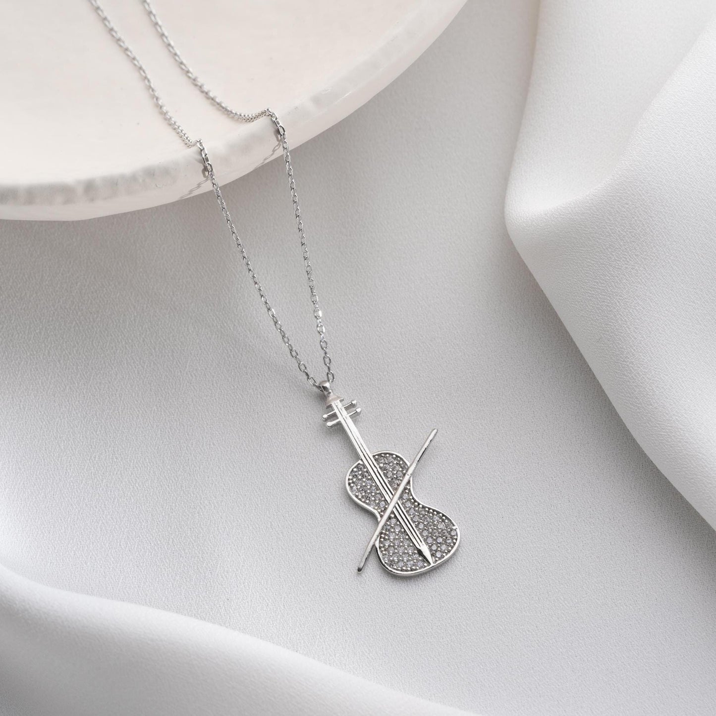 Violin-shaped necklace with cubic zirconia stones, available in 925 sterling silver and 14K gold plated options, displayed on a white satin background.