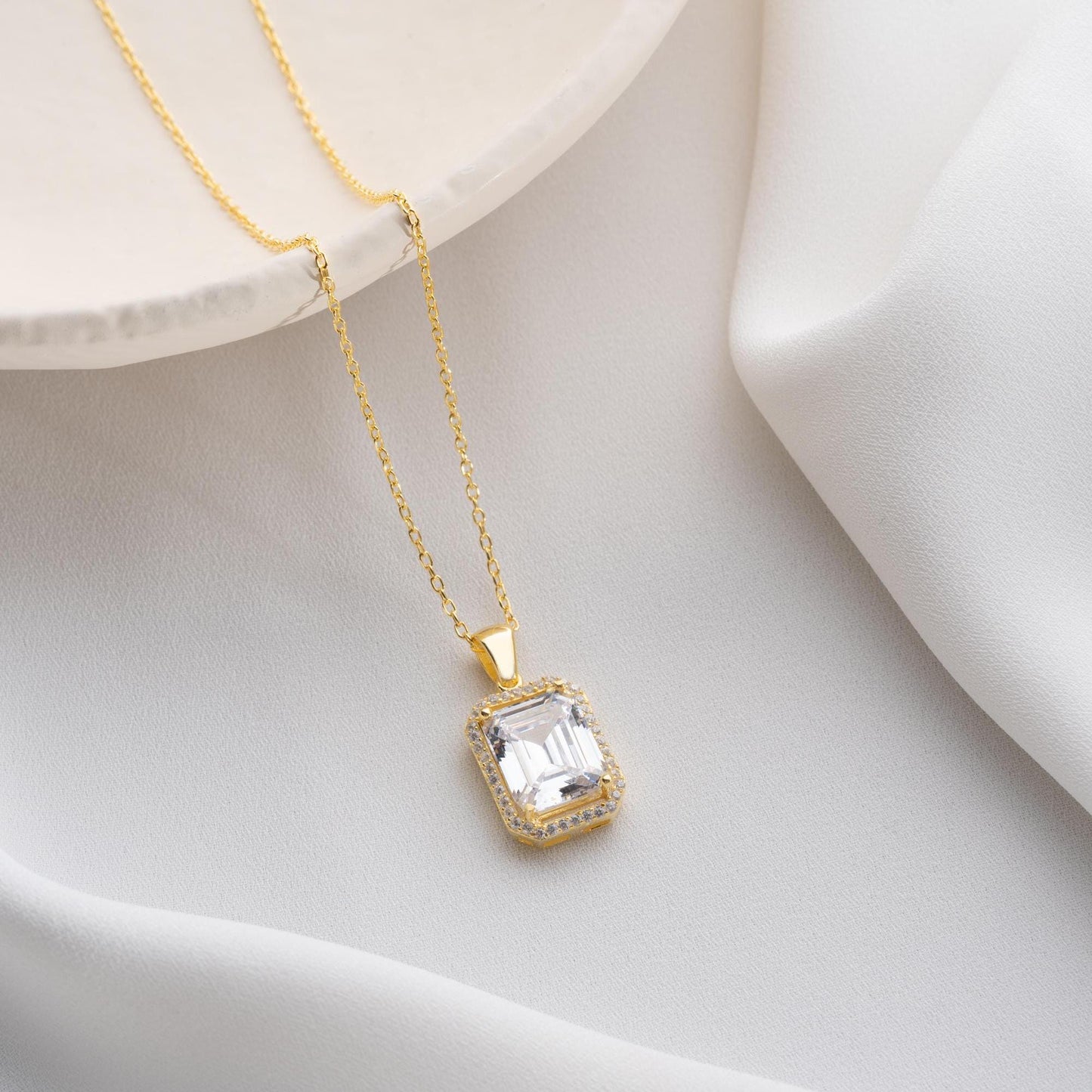 Emerald-cut pendant necklace with cubic zirconia stones, available in 925 sterling silver and 14K gold plated options, displayed on a white satin background