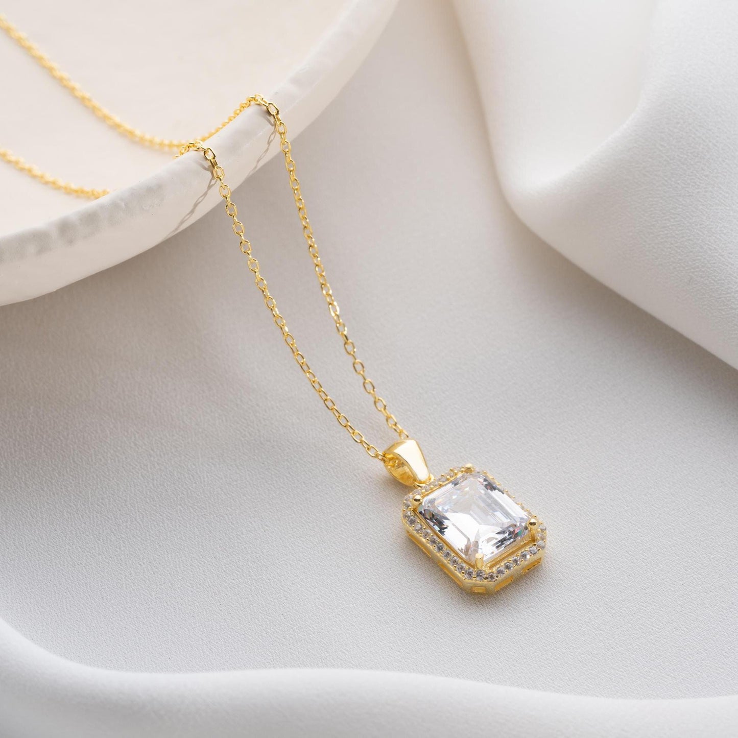 Emerald-cut pendant necklace with cubic zirconia stones, available in 925 sterling silver and 14K gold plated options, displayed on a white satin background