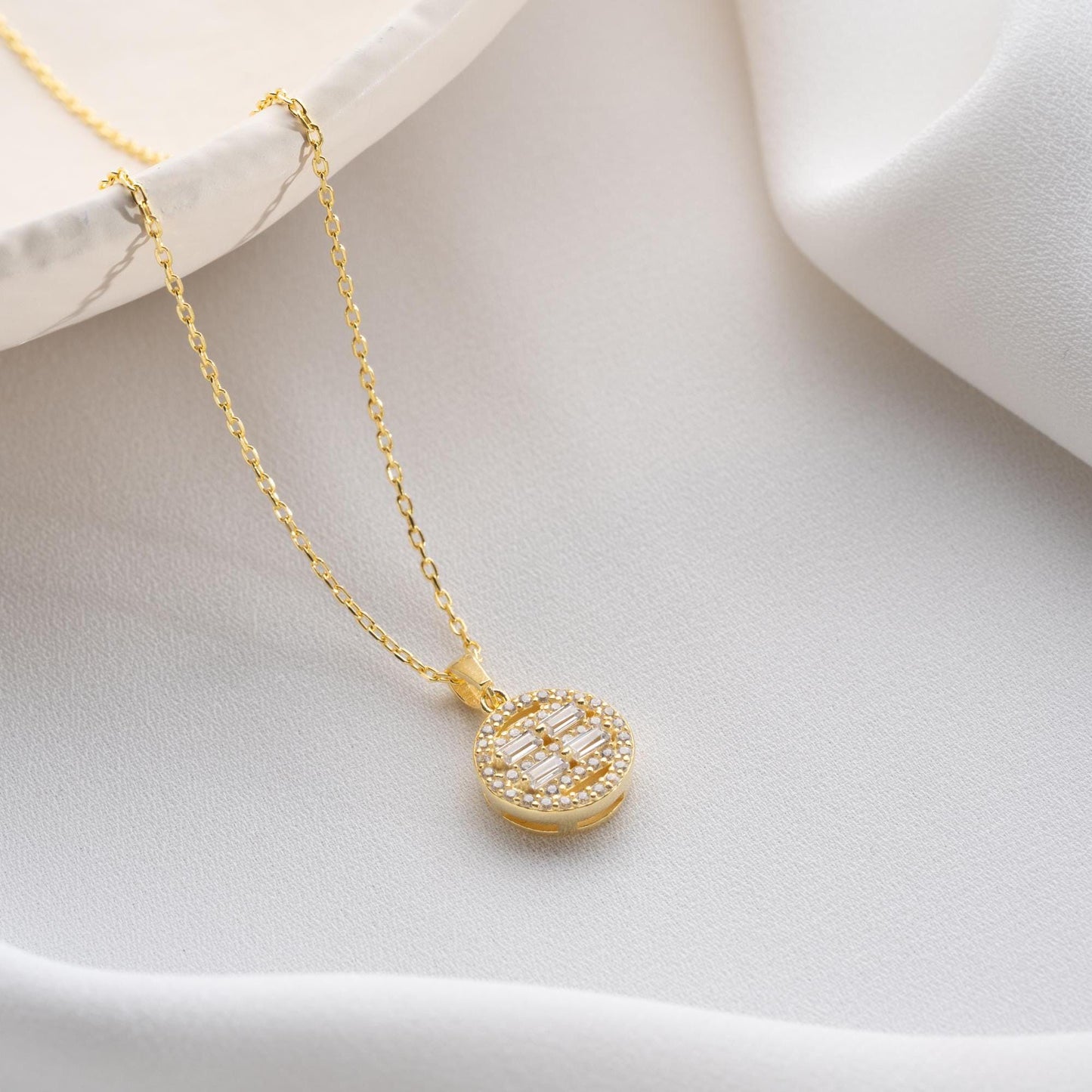 Round pendant necklace with cubic zirconia stones, available in 925 sterling silver and 14K gold plated options, displayed on a white satin background.