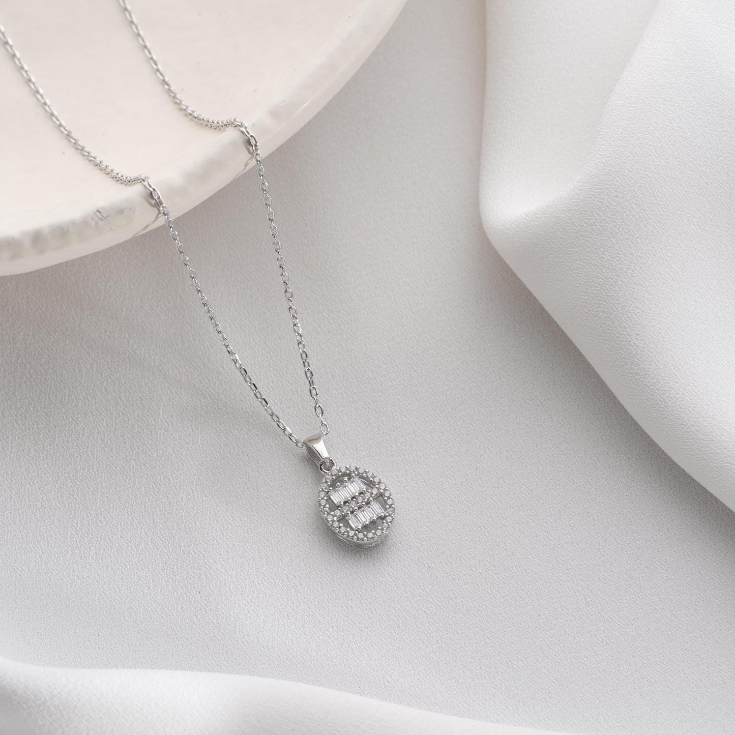 Oval pendant necklace with cubic zirconia stones, available in 925 sterling silver and 14K gold plated options, displayed on a white satin background