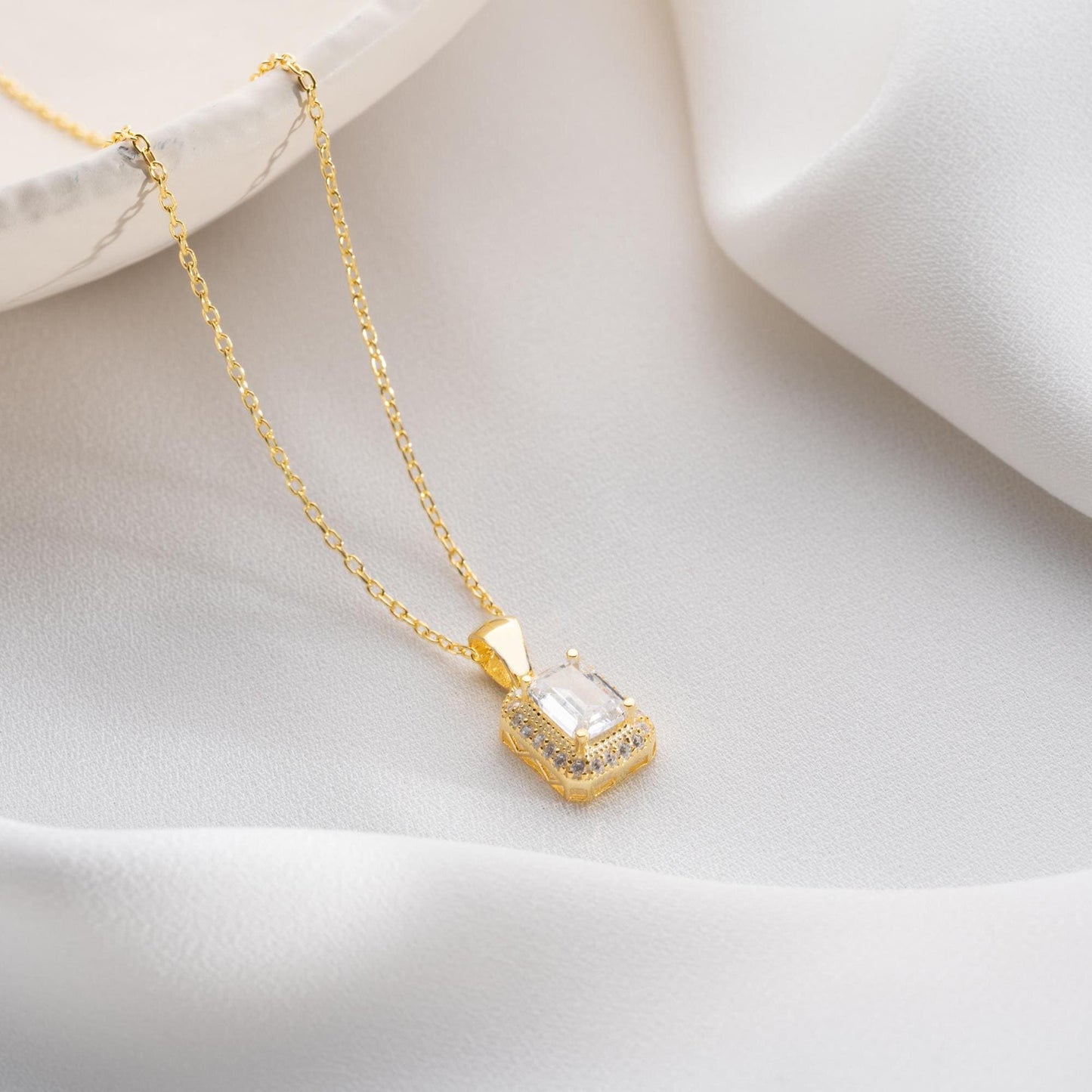 Emerald-cut pendant necklace with cubic zirconia stones, available in 925 sterling silver and 14K gold plated options, displayed on a white satin background.