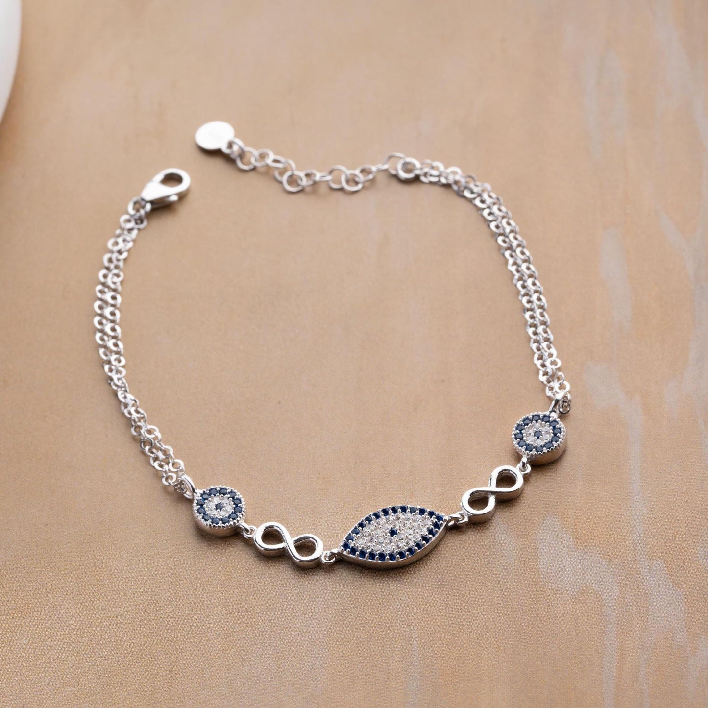 Elegant charm bracelet featuring evil eye and infinity symbols, crafted in 925 Sterling Silver with cubic zirconia stones, available in silver and gold finishes.