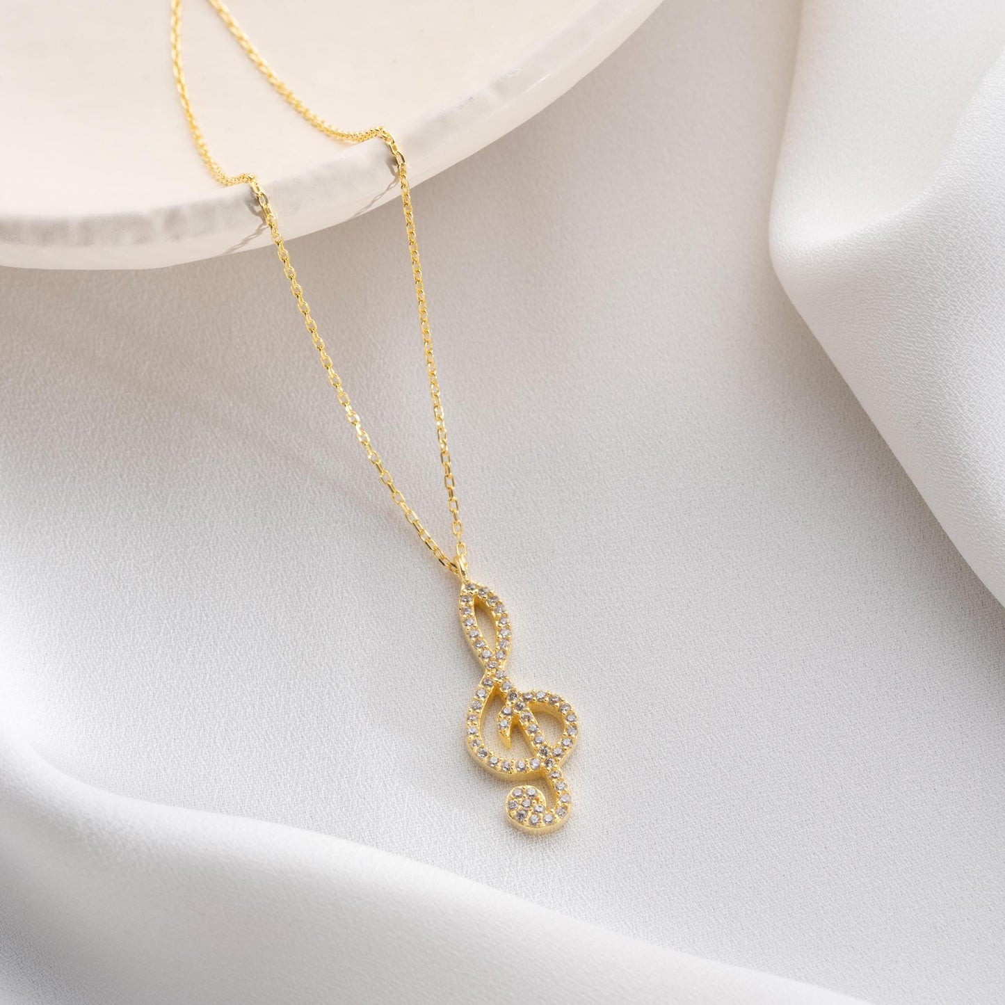 Music note necklace with cubic zirconia stones, available in 925 sterling silver and 14K gold plated options, displayed on a white satin background.