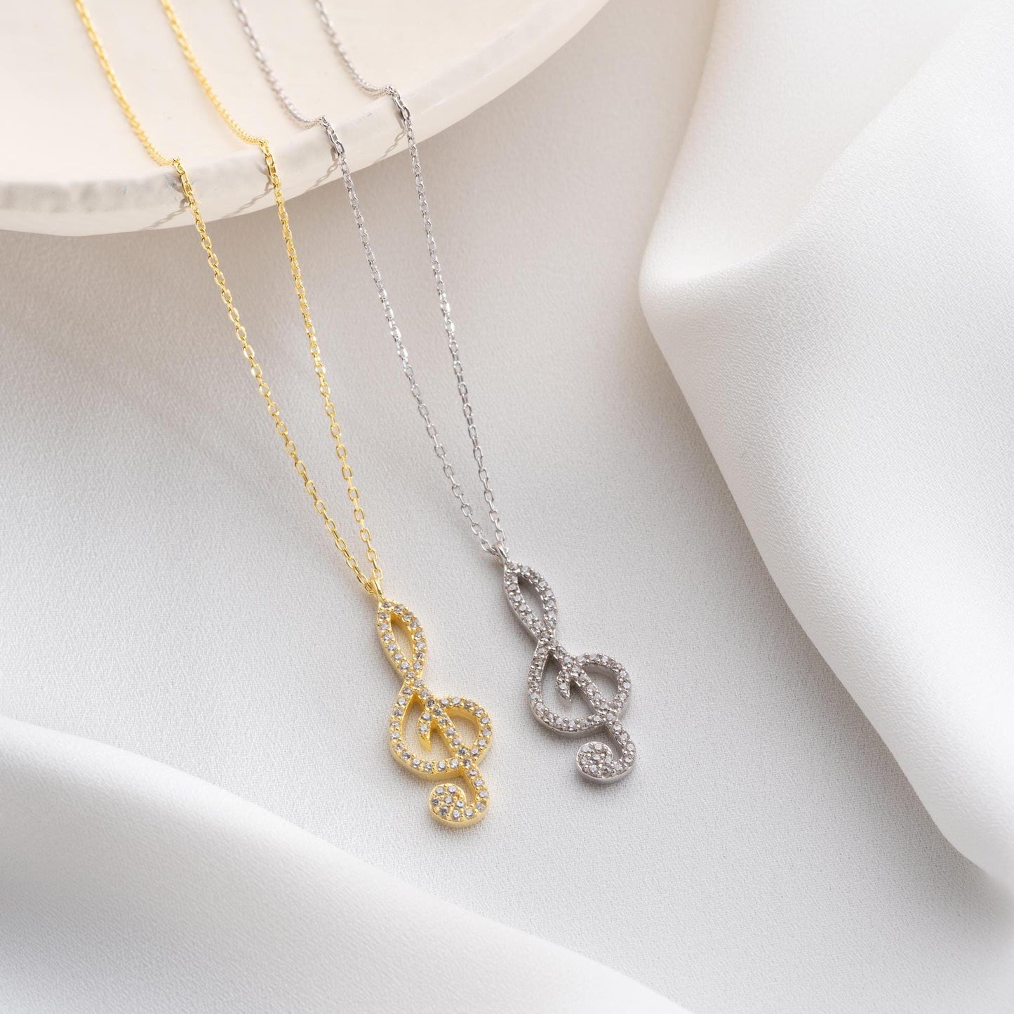 Music note necklace with cubic zirconia stones, available in 925 sterling silver and 14K gold plated options, displayed on a white satin background.