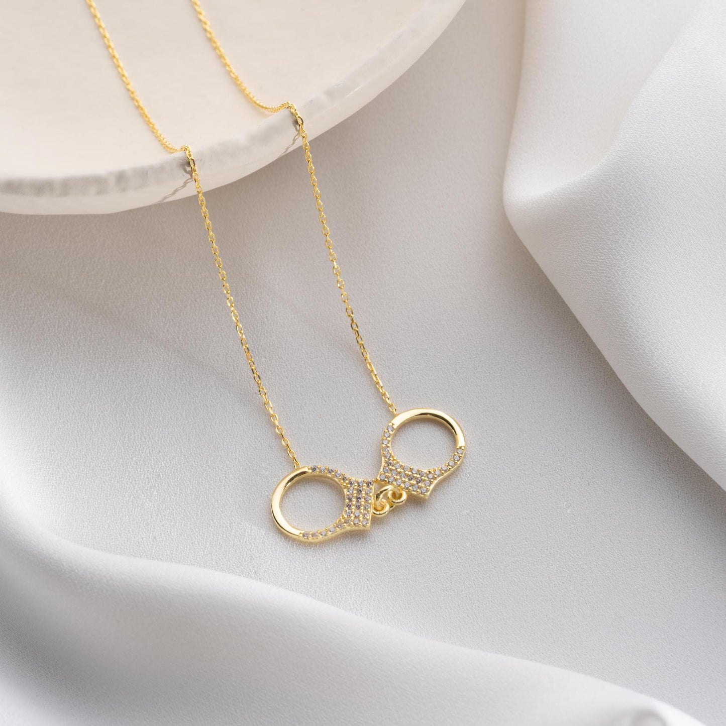 Handcuff necklace with cubic zirconia stones, available in 925 sterling silver and 14K gold plated options, displayed on a white satin background.