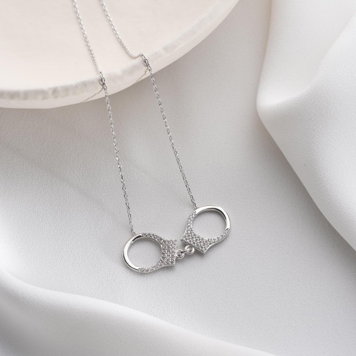 Handcuff necklace with cubic zirconia stones, available in 925 sterling silver and 14K gold plated options, displayed on a white satin background.