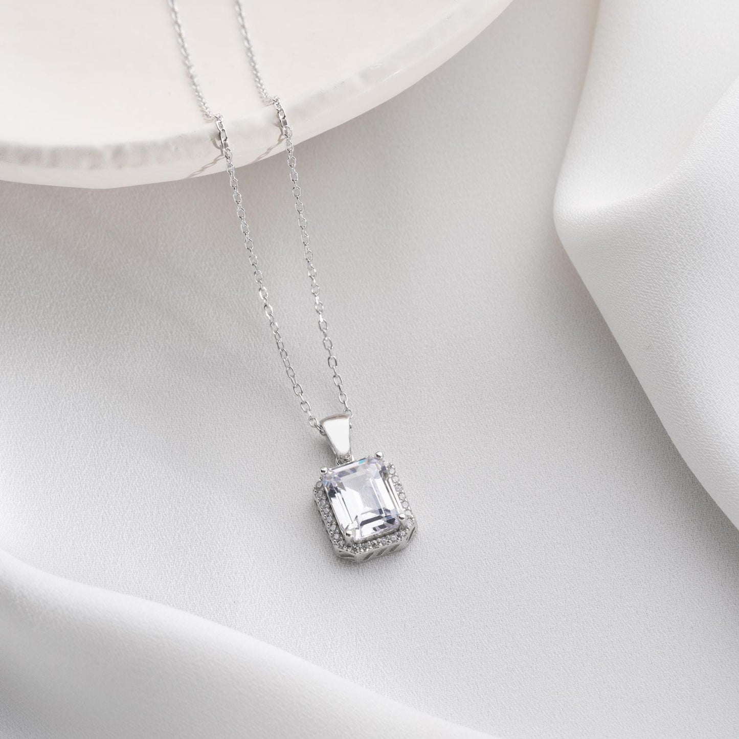 Emerald-cut pendant necklace with cubic zirconia stones, available in 925 sterling silver and 14K gold plated options, displayed on a white satin background.