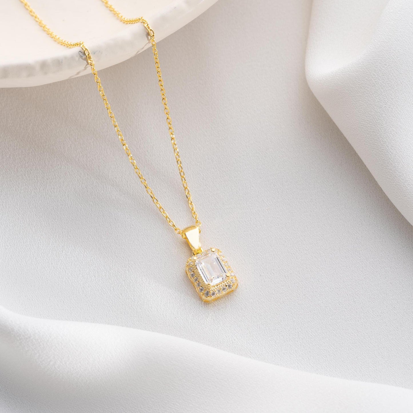 Emerald-cut pendant necklace with cubic zirconia stones, available in 925 sterling silver and 14K gold plated options, displayed on a white satin background.