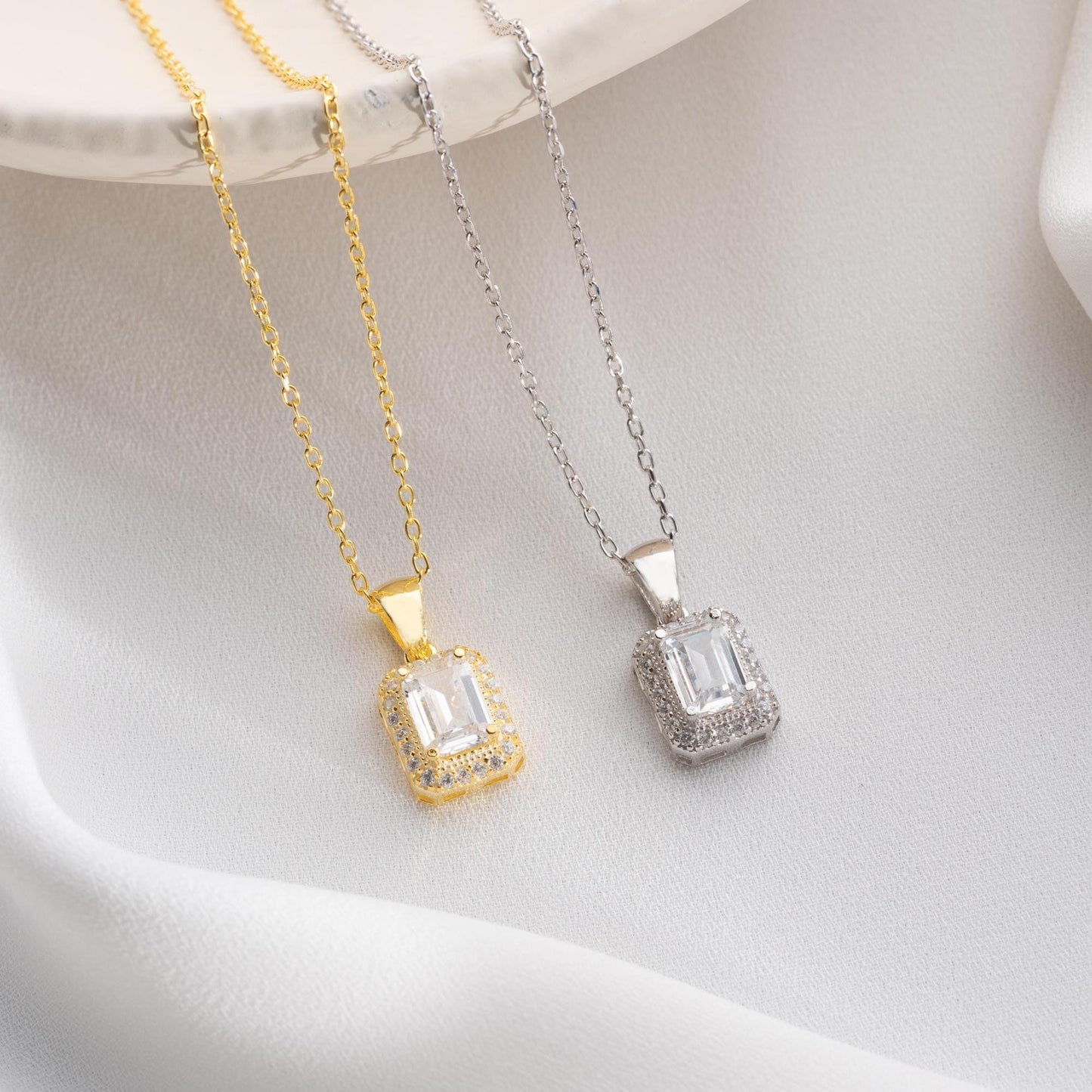 Emerald-cut pendant necklace with cubic zirconia stones, available in 925 sterling silver and 14K gold plated options, displayed on a white satin background.