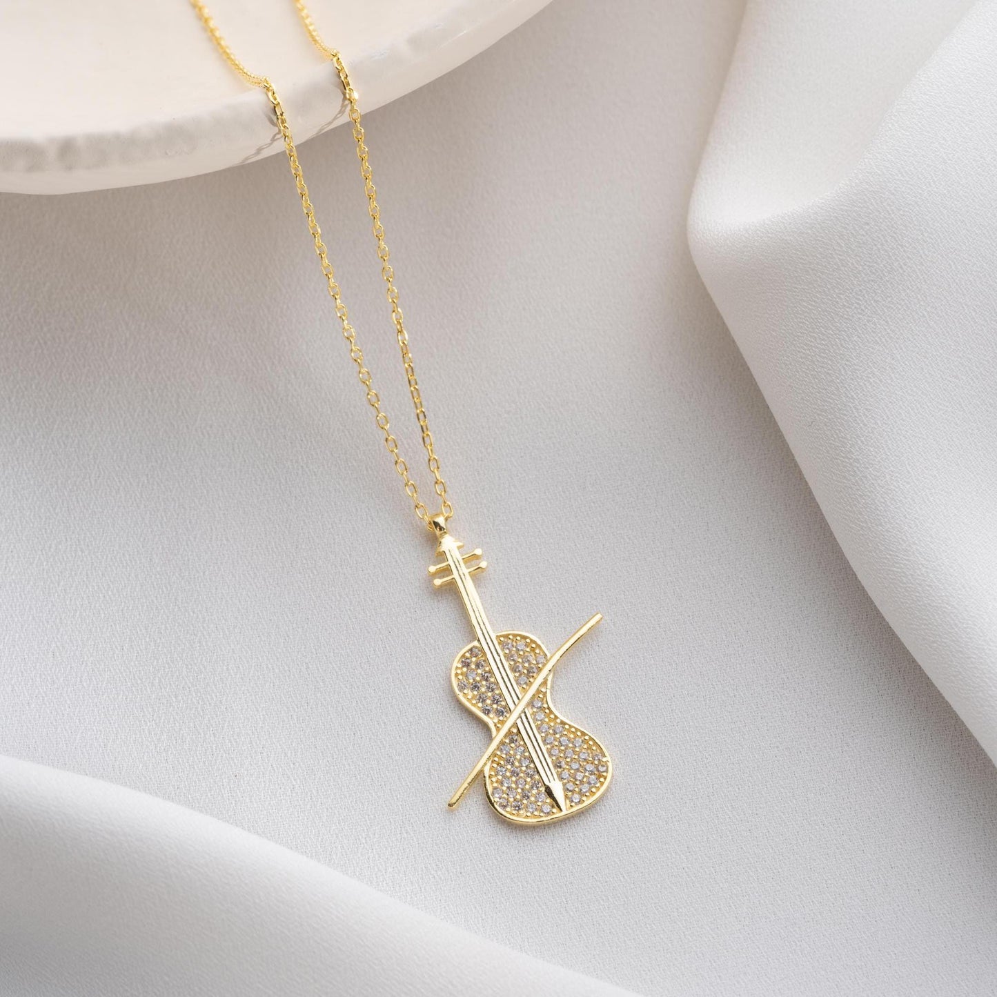 Violin-shaped necklace with cubic zirconia stones, available in 925 sterling silver and 14K gold plated options, displayed on a white satin background.