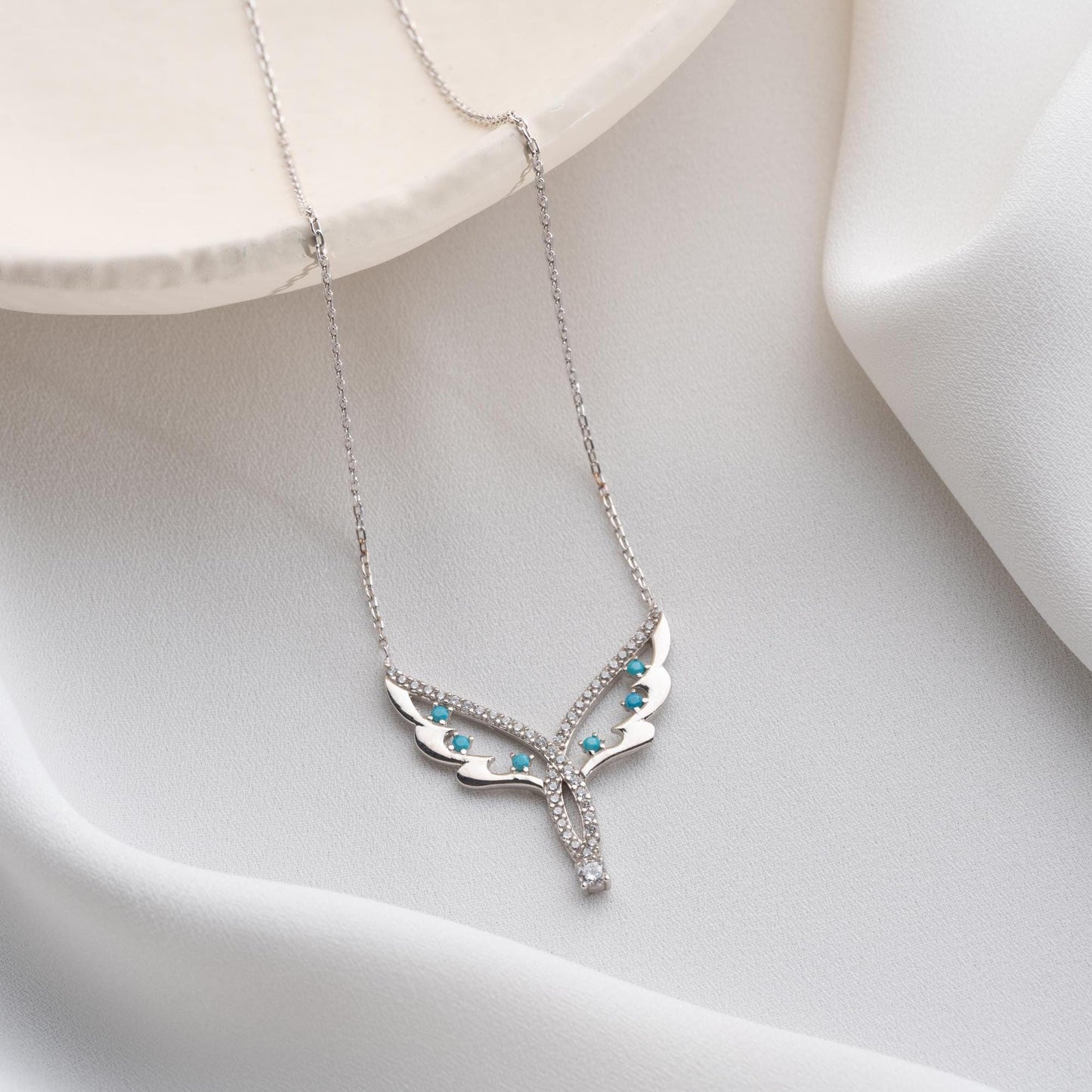 Angel wing necklace with turquoise and cubic zirconia stones, available in 925 sterling silver and 14K gold plated options, displayed on a white satin background