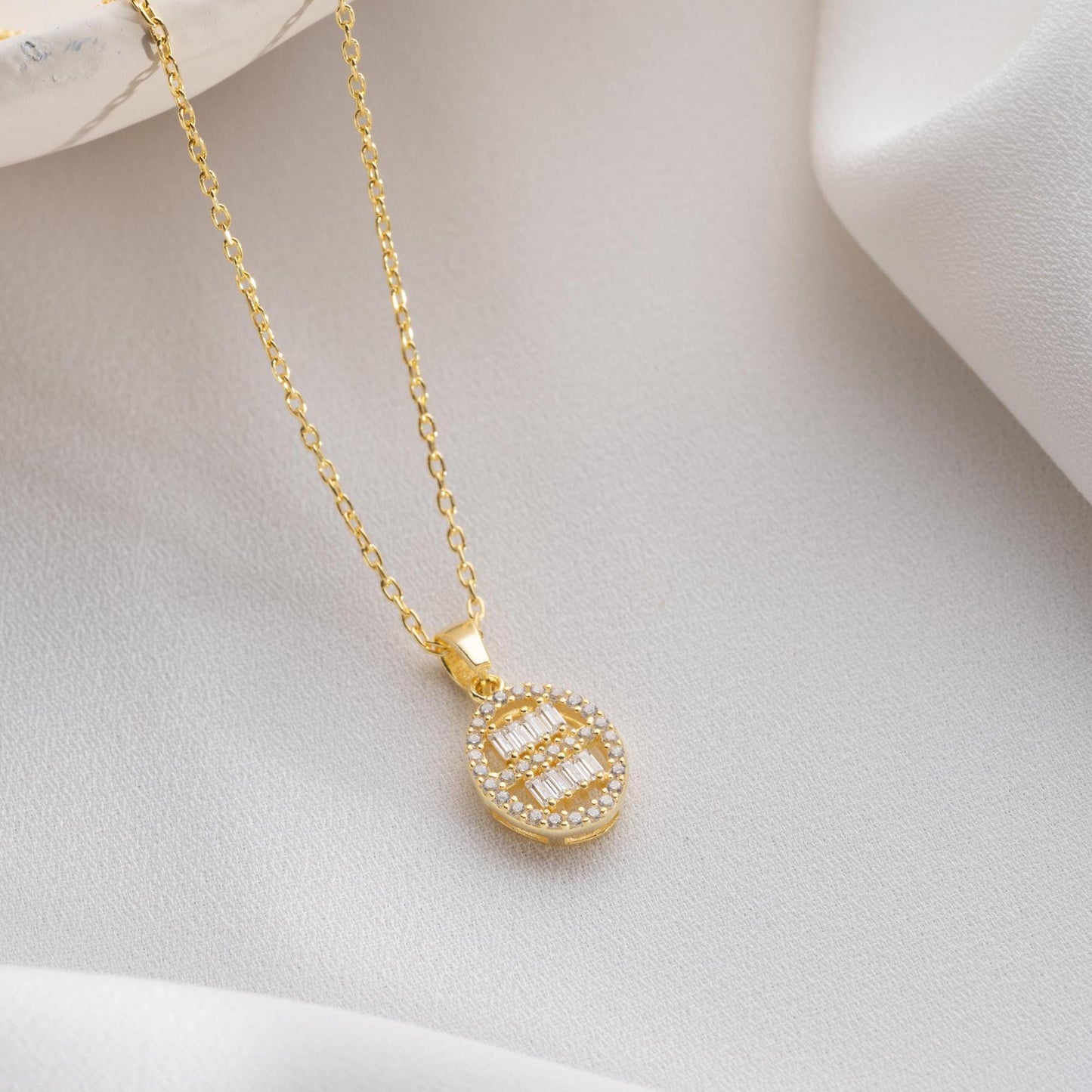 Oval pendant necklace with cubic zirconia stones, available in 925 sterling silver and 14K gold plated options, displayed on a white satin background