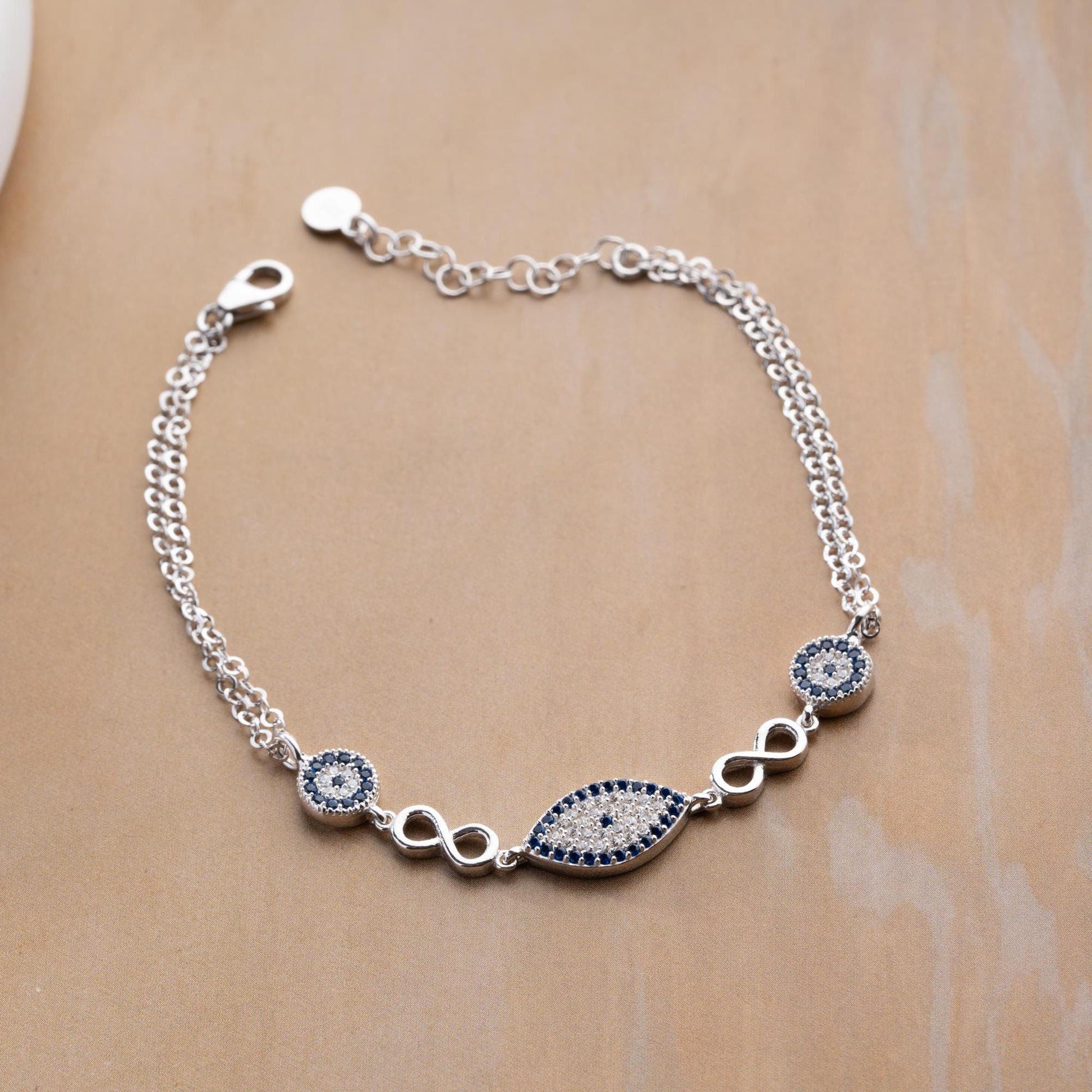 Elegant charm bracelet featuring evil eye and infinity symbols, crafted in 925 Sterling Silver with cubic zirconia stones, available in silver and gold finishes.