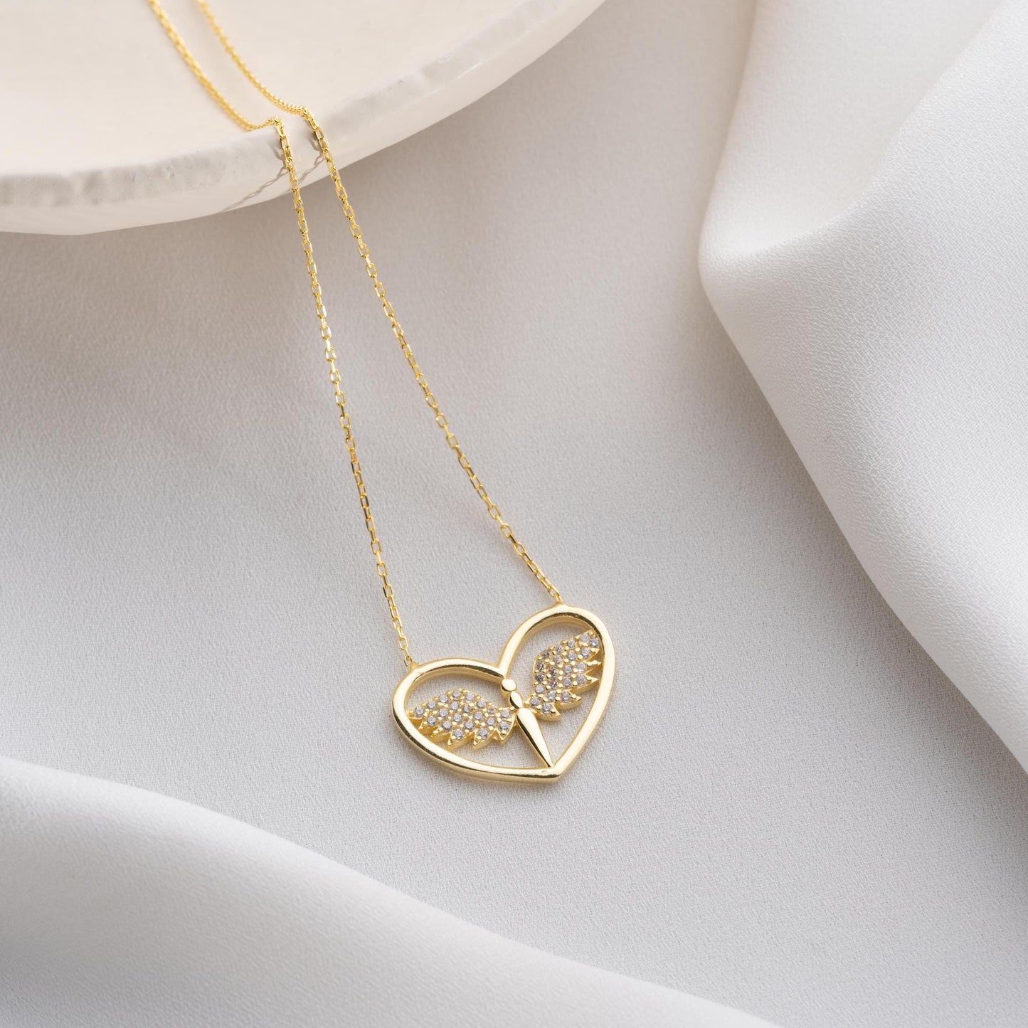 Angel wing heart necklace with cubic zirconia stones, available in 925 sterling silver and 14K gold plated options, displayed on a white satin background