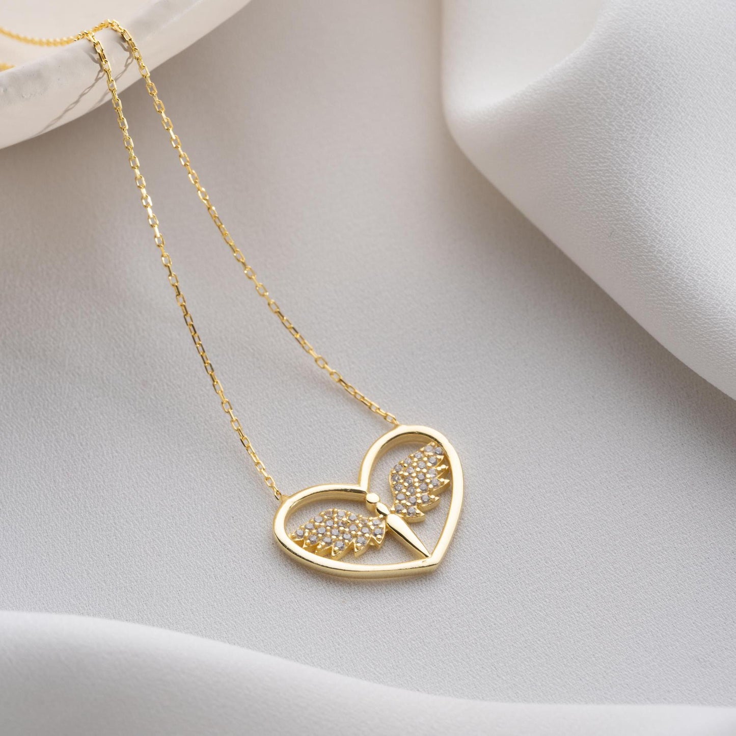 Angel wing heart necklace with cubic zirconia stones, available in 925 sterling silver and 14K gold plated options, displayed on a white satin background