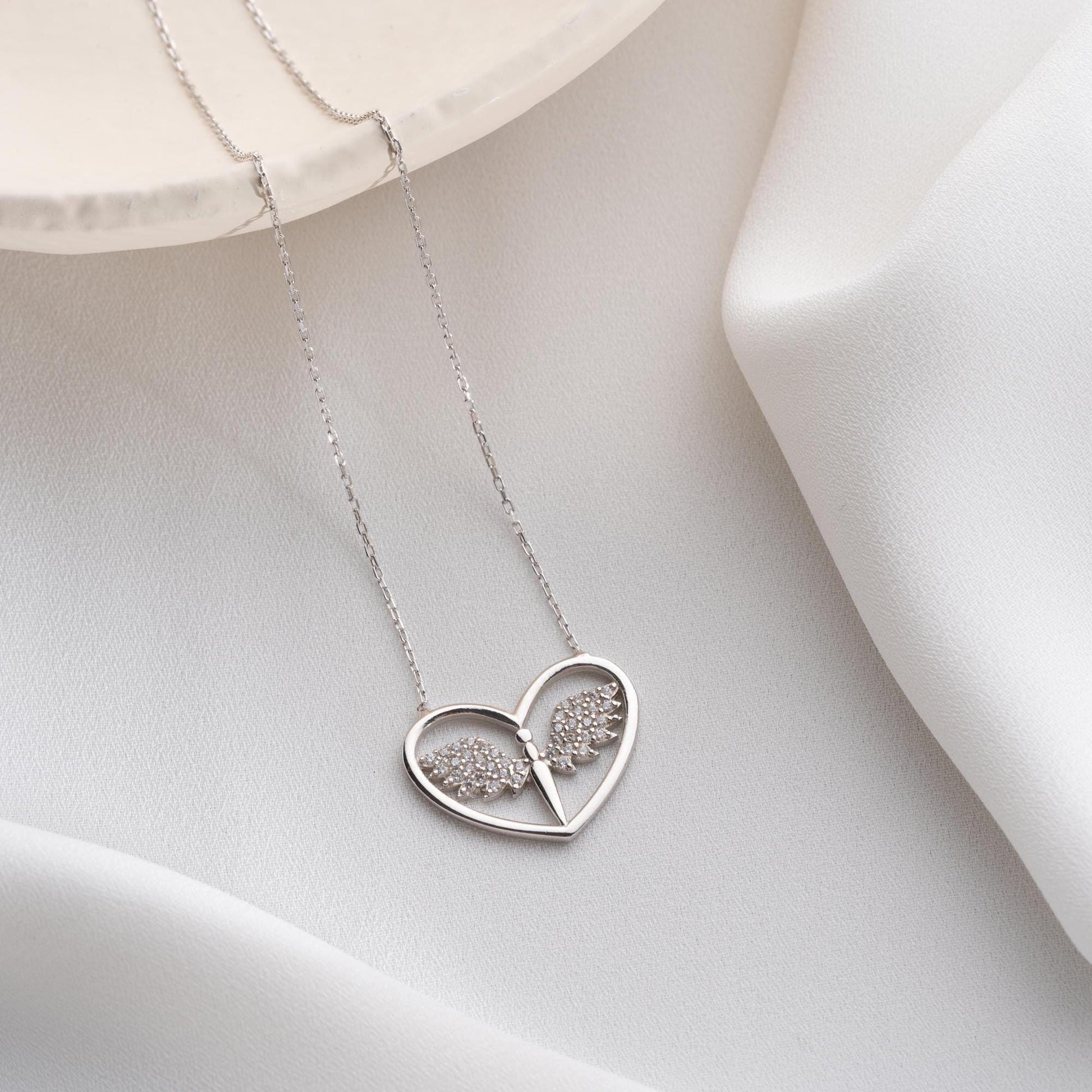 Angel wing heart necklace with cubic zirconia stones, available in 925 sterling silver and 14K gold plated options, displayed on a white satin background