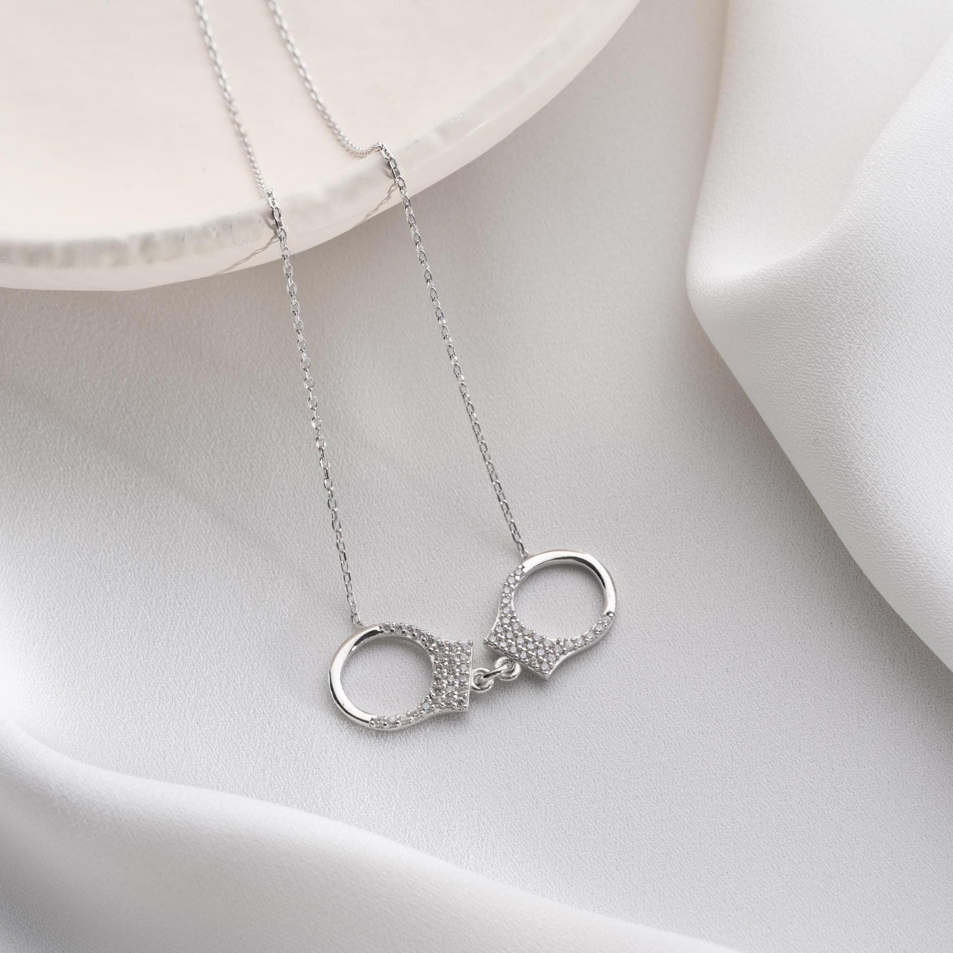 Handcuff necklace with cubic zirconia stones, available in 925 sterling silver and 14K gold plated options, displayed on a white satin background.