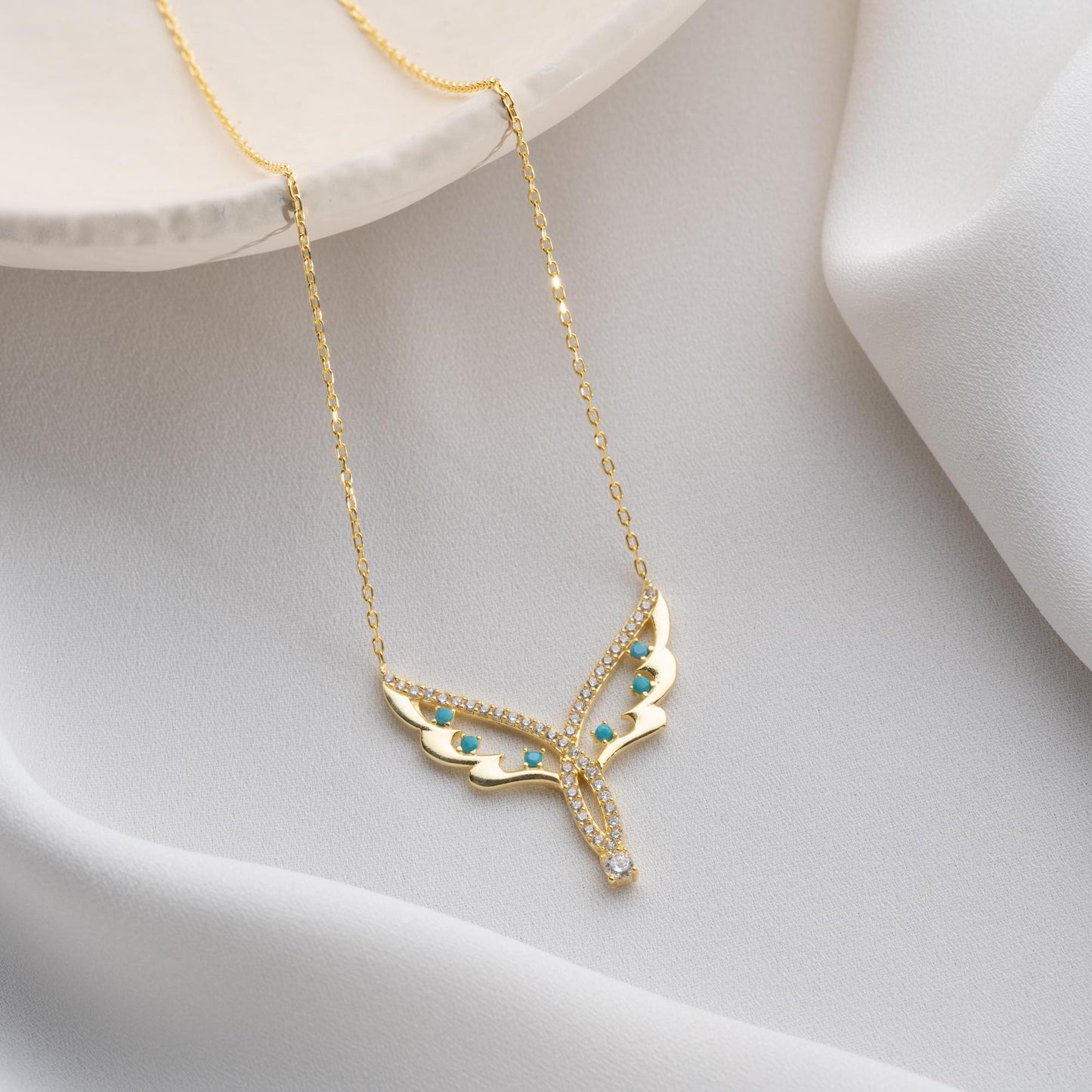Angel wing necklace with turquoise and cubic zirconia stones, available in 925 sterling silver and 14K gold plated options, displayed on a white satin background