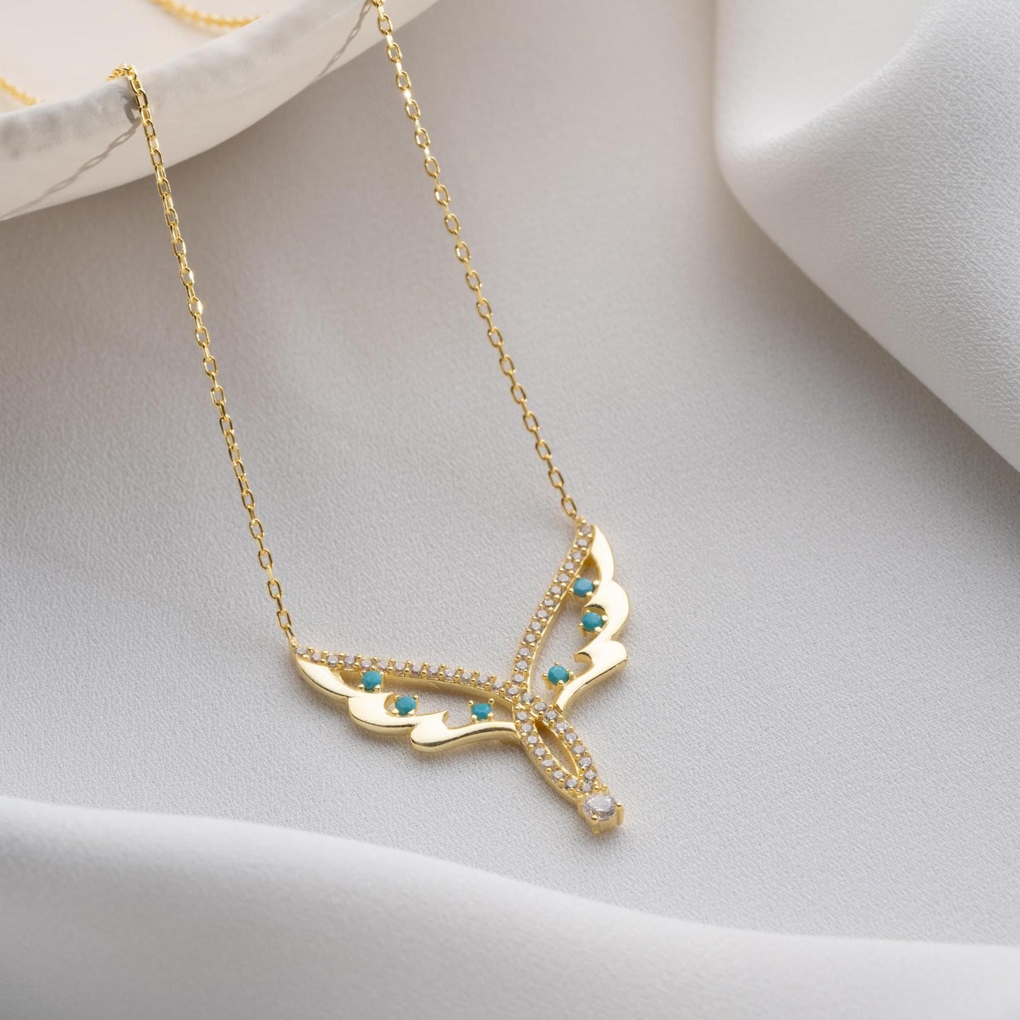 Angel wing necklace with turquoise and cubic zirconia stones, available in 925 sterling silver and 14K gold plated options, displayed on a white satin background