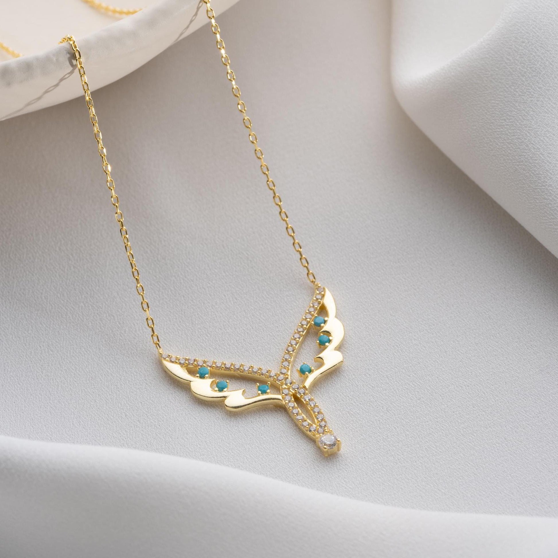 Angel wing necklace with turquoise and cubic zirconia stones, available in 925 sterling silver and 14K gold plated options, displayed on a white satin background