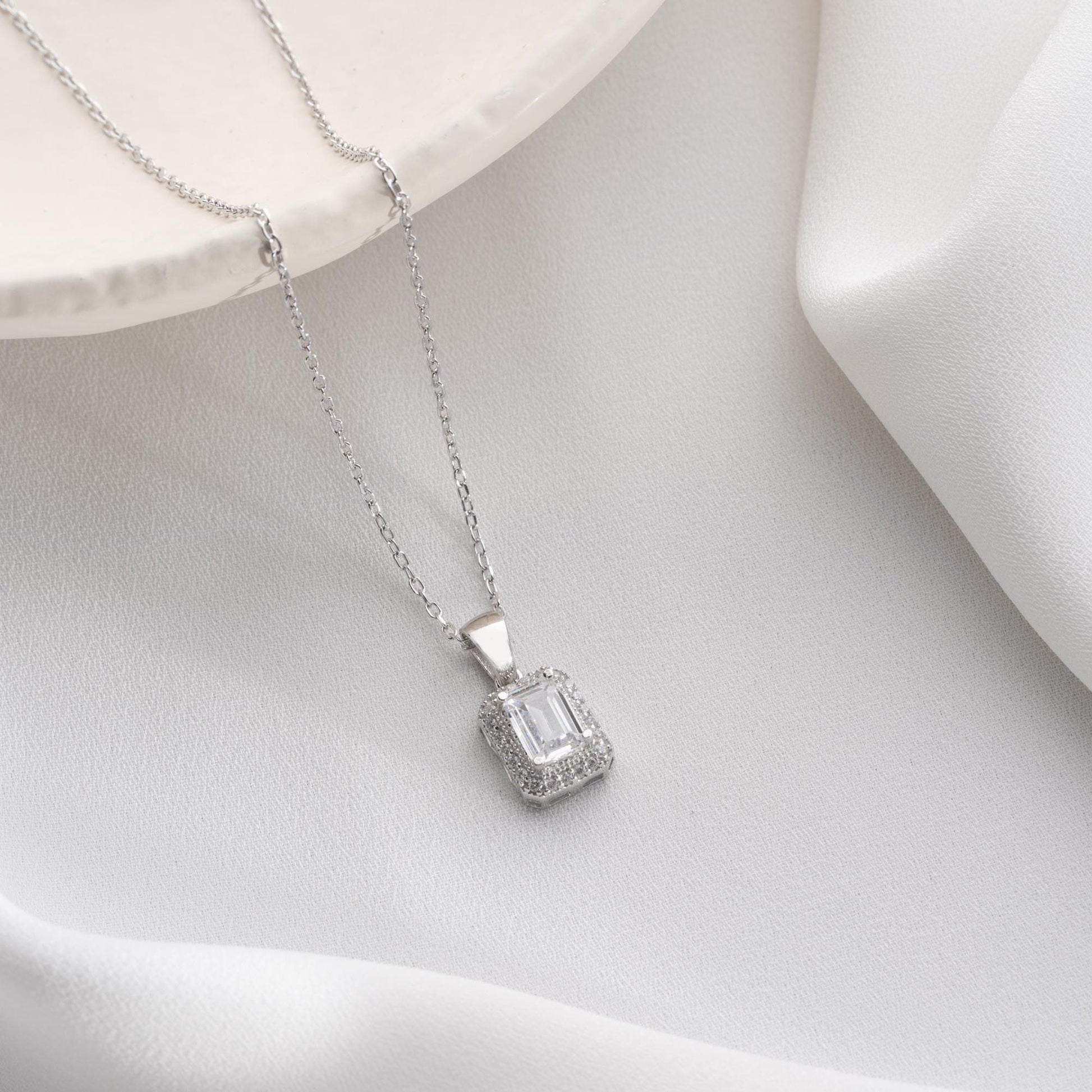 Emerald-cut pendant necklace with cubic zirconia stones, available in 925 sterling silver and 14K gold plated options, displayed on a white satin background.
